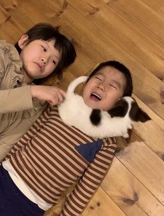 右側に寝ている男の子の首の上にマフラーの様に犬が乗っていて、左側にいる女の子が犬のしっぽを触っている写真
