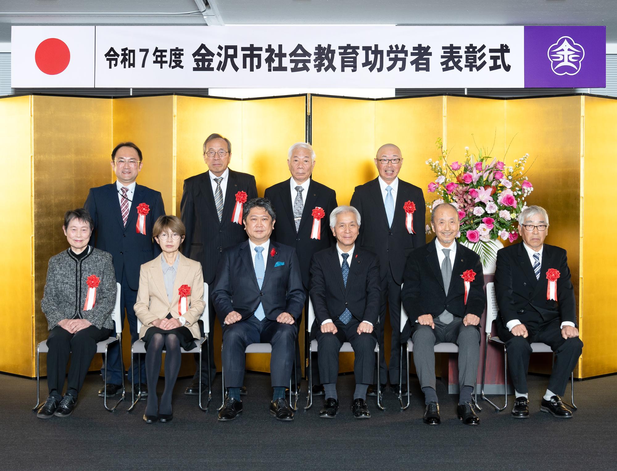 金屏風の前で胸に胸章を付けた受賞者たちが記念撮影をしている写真