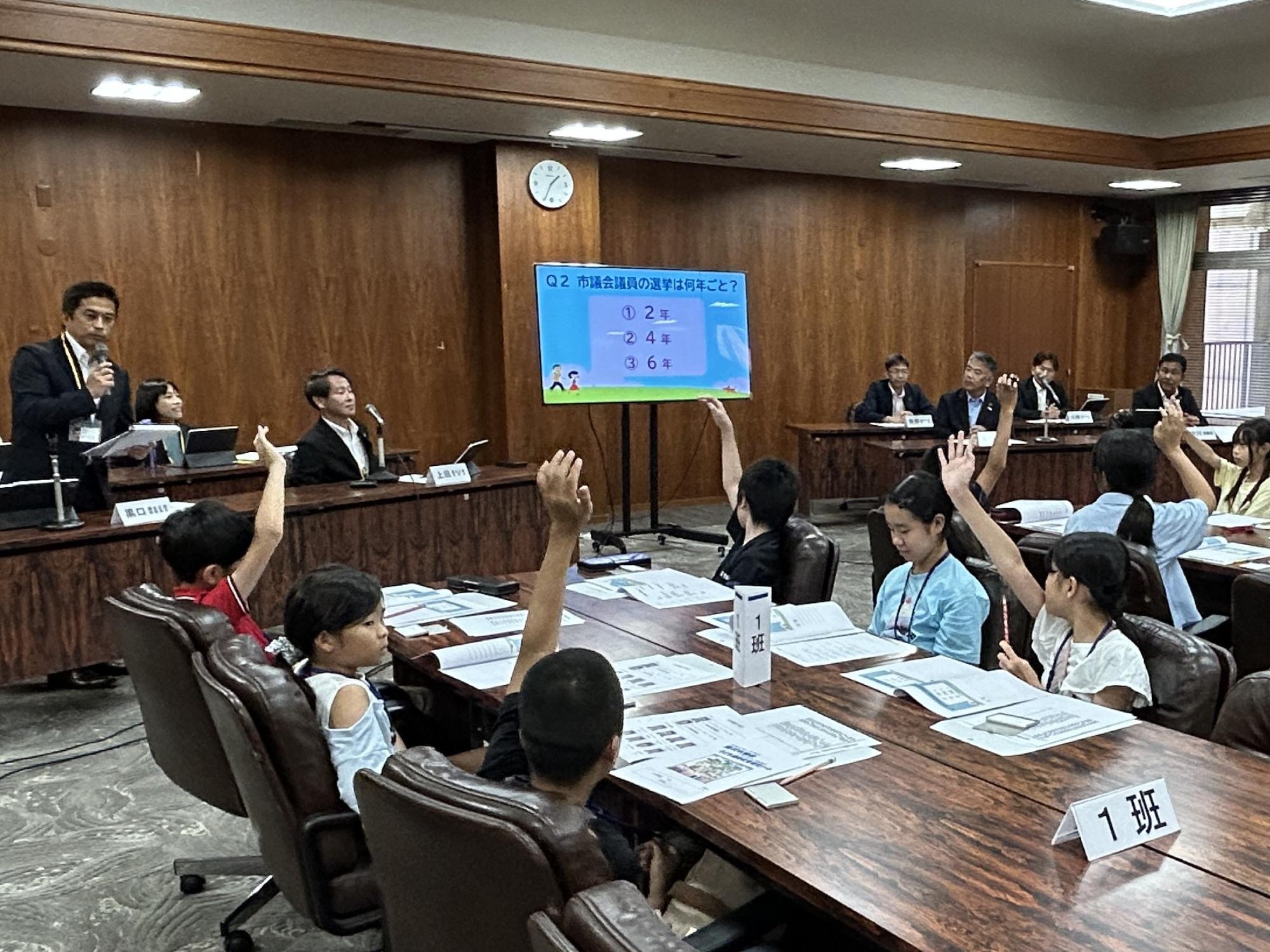 議会室内のスクリーンに資料が映し出され、席に座っている子ども達が説明を聞いている写真