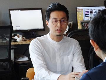 奥にパソコンが置かれている室内で柳井 友一氏と男性が向かい合って話をしている写真
