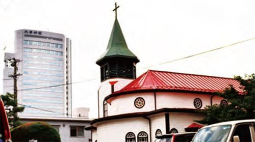 上に十字架のついた緑の鐘塔、下に赤の屋根がある白い建物が写っている金沢聖霊病院聖堂の外観写真