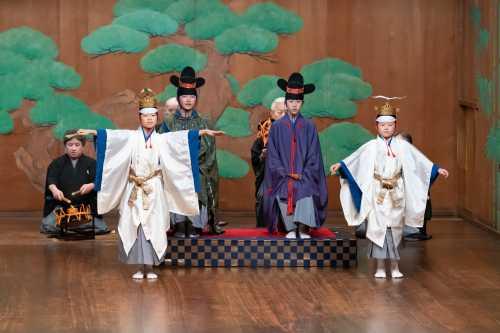 大きな松の絵が描かれた舞台で袴姿の大人たちが鼓をたたき、緑や紫色の能衣装を身に纏った男の子たちが立っており、その前で白い能衣装を身に纏った男の子たちが両手を広げて舞っている写真