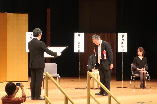 令和4年度金沢市文化活動賞の贈呈式のステージで胸に赤い花を付けた黒いスーツ姿の男性が表彰されているのを他の受賞者が椅子に座って見ている写真