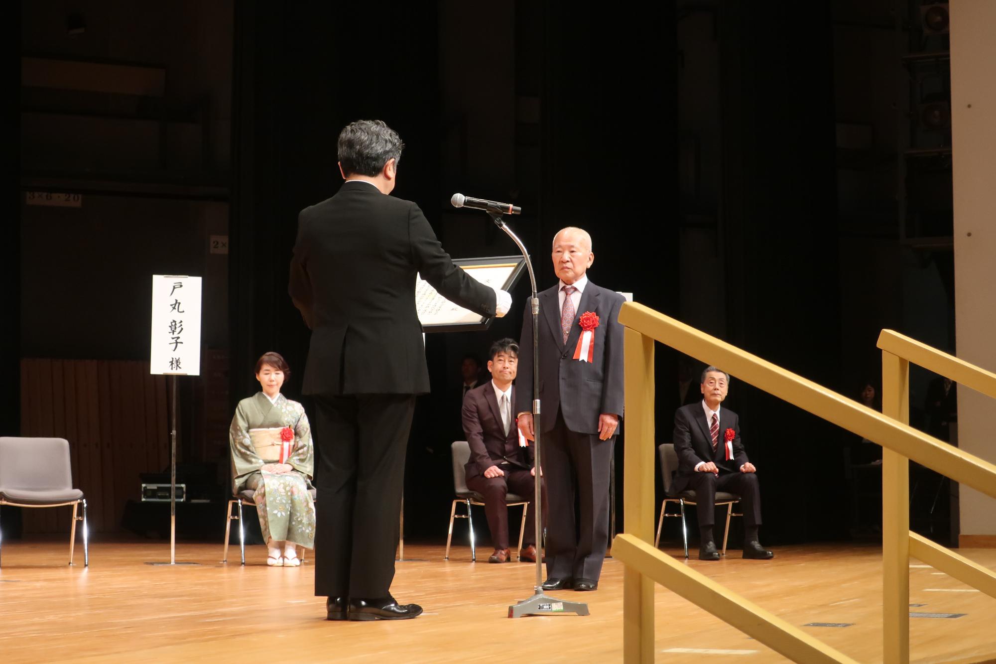 令和7年度金沢市文化活動賞の贈呈式のステージで赤い花を付けた男性が表彰されているのを他の受賞者が椅子に座って見ている写真