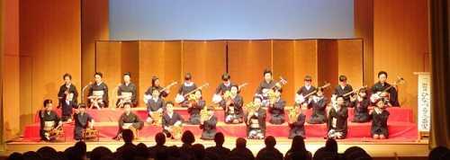 金屏風に赤いひな壇のステージで、黒い和服姿の女性たちが三味線、小鼓(こつづみ)、大鼓(おおかわ)、太鼓、笛のパートに分かれて演奏している写真