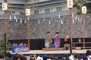 お城の前にある四隅に提灯が飾られている舞台に緑や紫色の能衣装を身に纏った男の子たちが立っており、後ろの方で袴姿の大人たちが鼓をたたき、舞台の左側には青い能衣装姿の子供たちが座っている写真