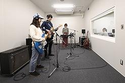 レコーディングスタジオでヘッドホンをつけた人たちがギターやキーボードなどで演奏している写真