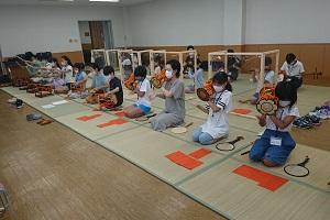 前列に小鼓(こつづみ)、大鼓(おおかわ)、太鼓を演奏する子供たちと、仕切りを付けた後列で笛を演奏する子供たちが合奏している写真