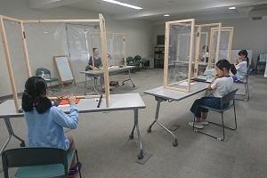 一人ずつ仕切りのされたテーブルに座り、笛の練習をしている子供たちを前のテーブルに座っている先生が見ている写真