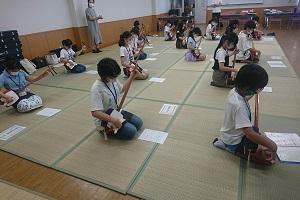 畳に座り楽譜を見ながら三味線を演奏する子供たちを後ろの方で先生が立って見ている写真