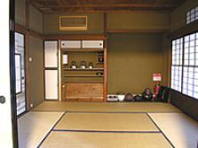 入口が障子戸の和室の床の間に茶道具が置いてあり左隣に棚がある茶室の写真