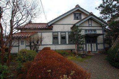 建物の周囲に植えられた樹木や生垣がある、赤茶色の屋根に白い外壁の飯田家住宅の外観写真