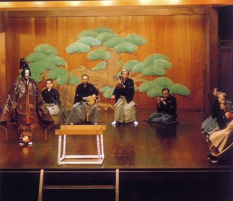 舞台で能面をつけて能の衣装を身に纏った人が舞をしている後ろで、袴姿の男性たちが太鼓や笛を演奏しており、右側には袴姿の男性たちが座っている加賀宝生の写真