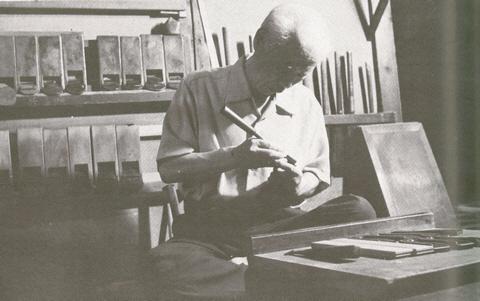 かんななどの指物製作用具が棚に並んでいる前で道具を手に持ち指物製作をする男性の白黒写真