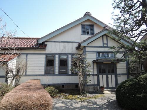 赤い瓦の三角屋根の建物で、外壁が白で、窓枠や入口が濃いグレーの枠の飯田家住宅主屋の外観写真