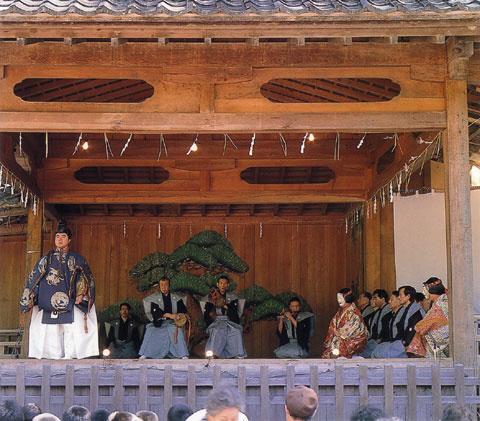 大野湊神社の舞台の前方左側に、能の衣装を着た男性が立ち、後ろの方で鼓や笛の演奏が行われており、右側では能面を付けた人や袴姿の人々が座っている寺中神事能の写真