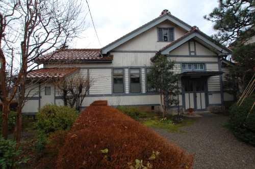 建物の周囲に植えられた樹木や紅葉した生垣、赤茶色の屋根に白い外壁の飯田家住宅の写真