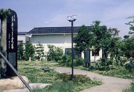 木々の植えられたチカモリ遺跡公園奥にある白い壁の金沢市埋蔵文化財収蔵庫の外観写真