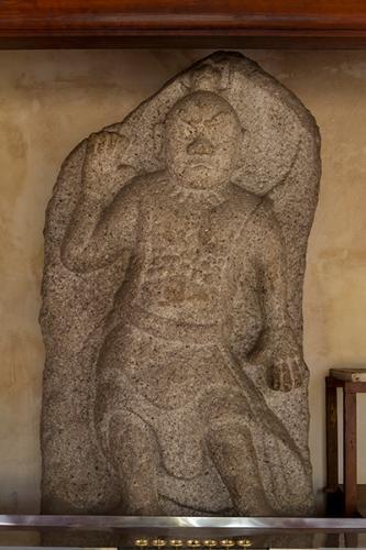 肘を曲げて右手を挙げ、左手は下におろして立っている、石の表面に掘られた石造金剛力士立像（吽形）の写真