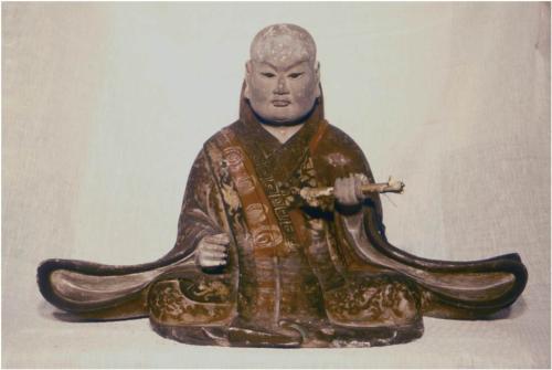 衣の上に袈裟をかけ、左手に経巻を持ってあぐらを組んで座っている木造日蓮聖人坐像の写真