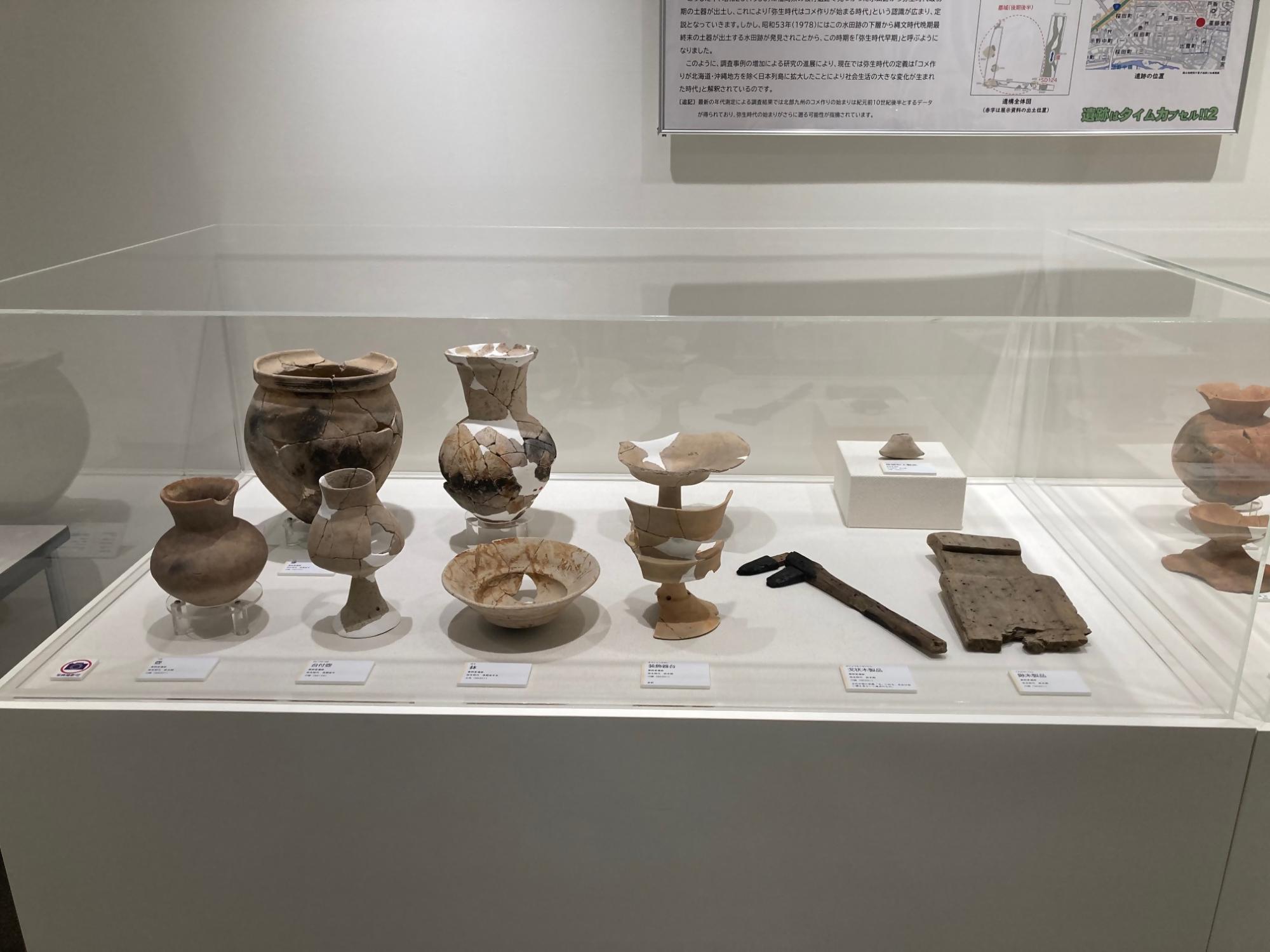 弥生時代の土器や木製品が展示台に並んでいる企画展「遺跡はタイムカプセル!!2」の展示風景