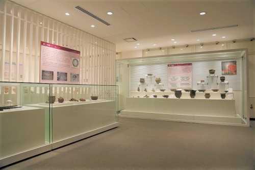 左側には説明の書かれたパネルが展示され、その下に出土された土器や石器が並べられカラスケース、奥には大きなガラスケースに出土された土器や石器が2段になっている台の上と壁に着けられた台に展示されている常設展示の写真