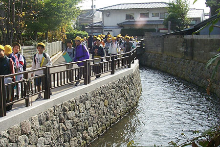 水が流れている用水路の左側の歩道に、黄色の帽子を被った子供が列をなして歩いている写真