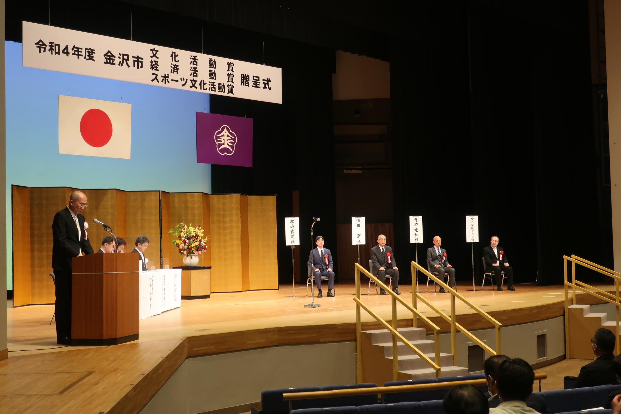 壇上の左側で山田副市長が選考結果を報告しており、右側に受賞者が着席している
