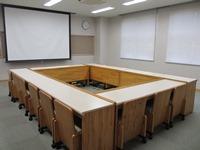 木製の机と椅子がロの字に設置され、大きなスクリーンがある会議室の写真