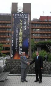 山出市長と福光実行委員長が時計台に掲げられた「ユネスコ クラフト創造都市に認定」と書かれている懸垂幕の前で握手をしている写真