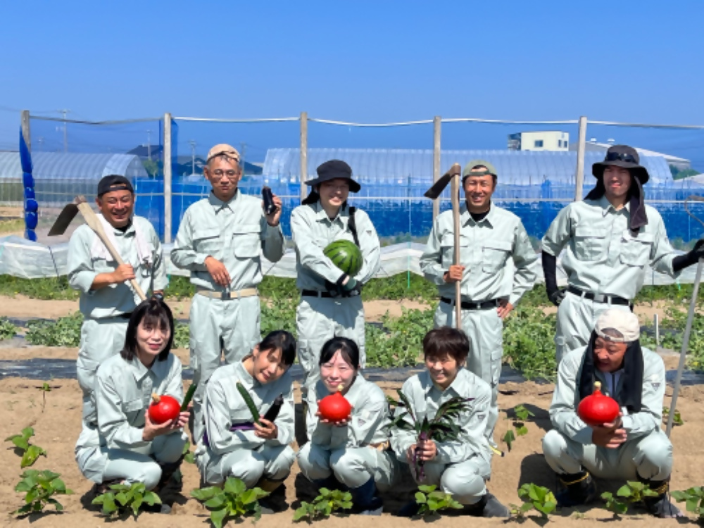 作業着を着た10名の研修生が、野菜の植えられた畑で、採れた野菜を持って笑顔で写っている写真