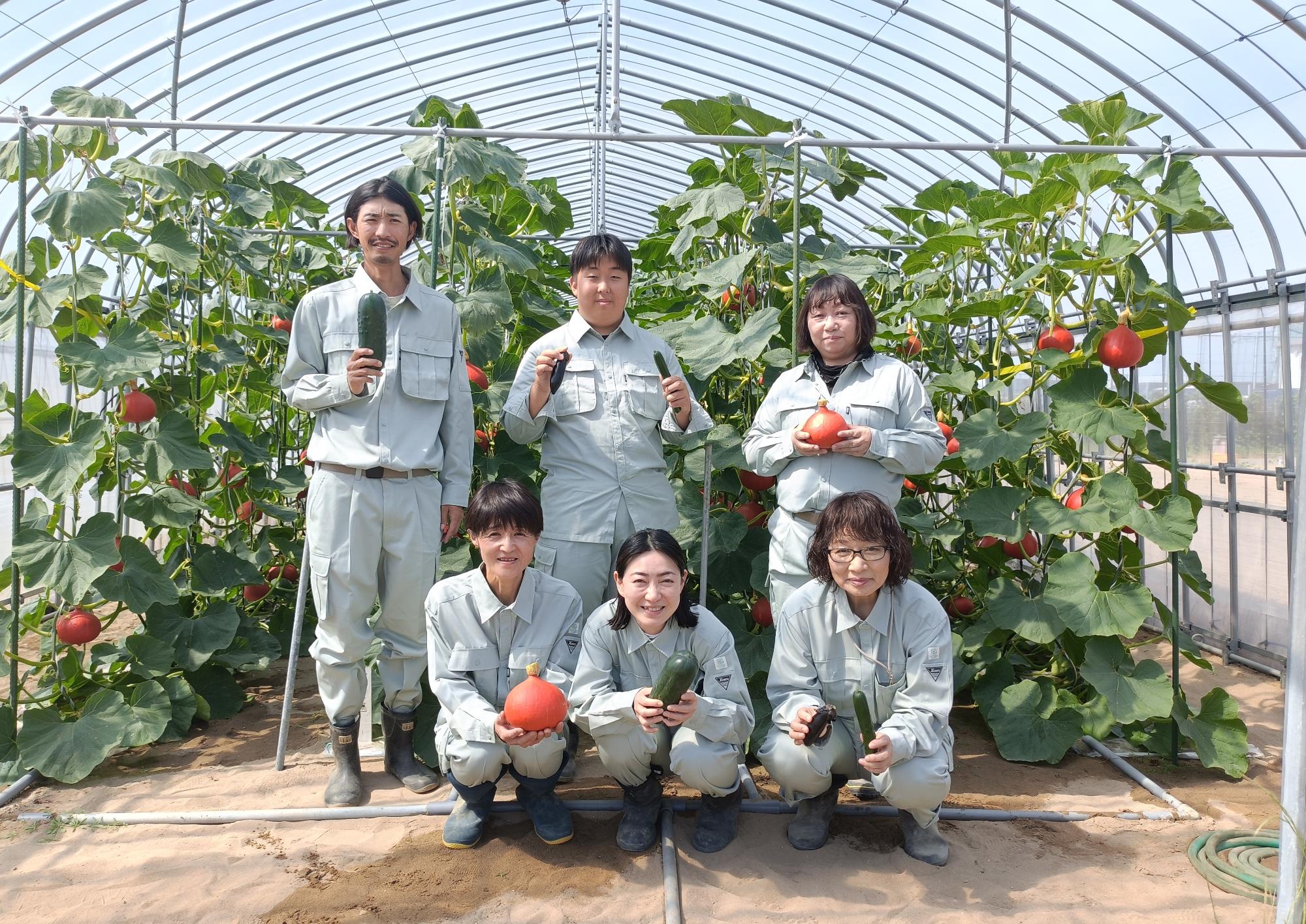 作業着を着た研修生が、野菜の植えられた畑で、採れた野菜を持って笑顔で写っている写真