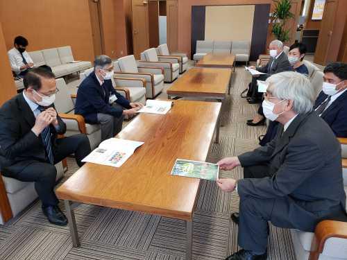 市長や関係者の方々が向かい合ってソファーに座っており、白髪の男性が話をしている、市長と左隣に座っている男性が木製のテーブルに置かれた資料を見ている様子の写真