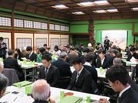 机の上に緑の札をたて、スーツを着だ男性がグループごとに着席している写真
