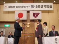 日本国旗がかけられた舞台上で協同組合金沢問屋センターの関係者が向かい合って立っている市の関係者と宣言書の受け渡しをしている写真