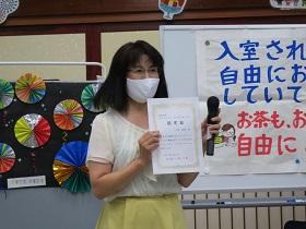 認定書を正面に向けて持ち、マスクを付けた状態でマイクを持ち抱負を述べている石黒佳恵さんの写真