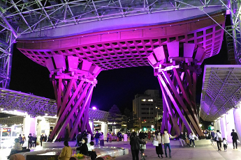 老若男女が大勢歩いている金沢駅鼓門の周辺が紫色でライトアップされている写真