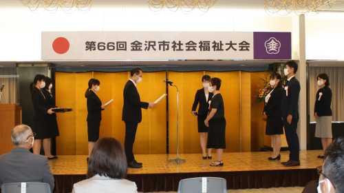 第66回金沢市社会福祉大会と書かれた横断幕の下の壇上で、スーツを着た男性がマイクの反対側に立っている女性を表彰している贈呈式の様子の写真