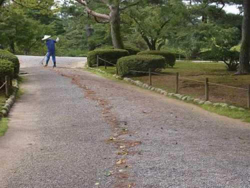 園の歩道の落ち葉をほうきでかき集めて掃除をしている男性の後ろ姿が歩道の奥に写っている写真