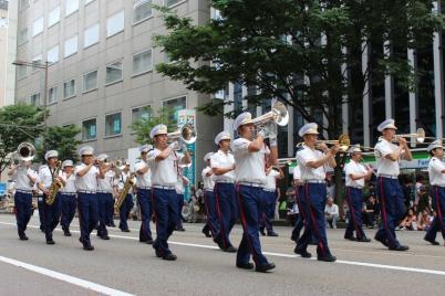 紺色のスラックスと白いシャツに身を包んだ消防音楽隊の人たちがトランペットを吹きながら観客に囲まれた道路を行進している写真