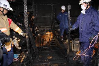 火災で真っ黒に燃え残った物を検分する消防隊員3人の写真