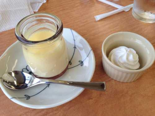 瓶に入った手作りプリンとスプーンが小皿の上に置かれ、白色の小さな器に生クリームが入っている写真