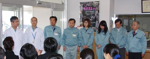 白衣を着た男性2名と作業着を着た6名の男女が関係者の前に立っている写真