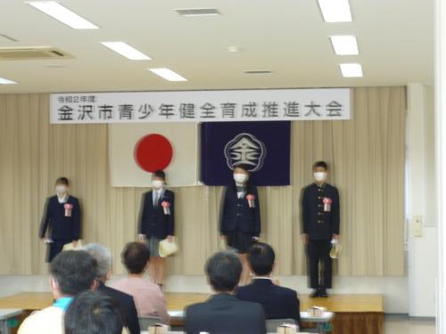 ステージ上で、胸章リボンをつけた4名の高校生が賞状を持ち、関係者の方を向いて立っている写真