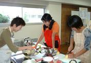 エプロンを付けた3名の女性が調理台で材料を切ったり、盛り付けたりしている写真