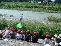 奥には川、草むらに立つ男性、手前には石段に座る生徒たちが写っている写真