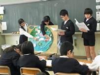 黒板の前に4人の生徒が立って、丸い絵の描かれた資料を使いながら説明をしている写真