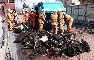 消防士たちがゴミ回収車の周りで作業していて、手前には炭のように焼けたゴミが置かれている写真
