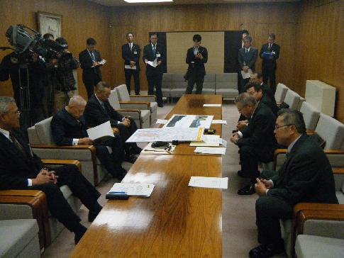 報道のカメラが入っている室内で、中央のテーブルを挟んで左側に男性3名、右側に男性4名が座っている写真