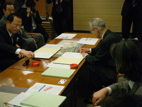 テーブルの左側に座っている市長と、右側に座っている関係者の男性がそれぞれの書類に判を付こうとしている写真
